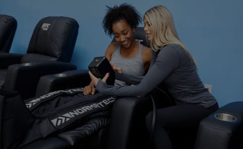 Two women sitting on a black massage chair against a blue background, looking at a device together